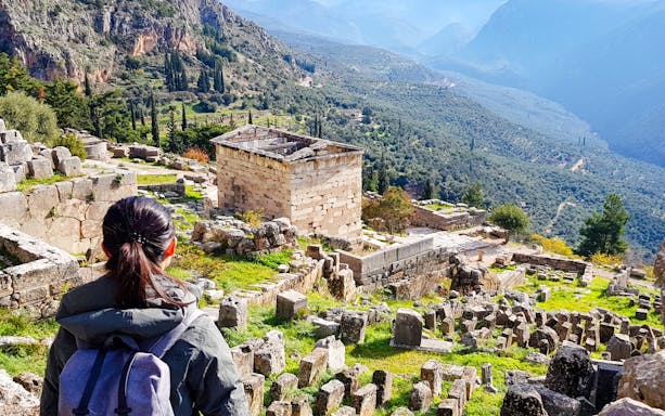 Visitor exploring ancient ruins at Delphi Archaeological Site, Greece, with mountainous landscape.
