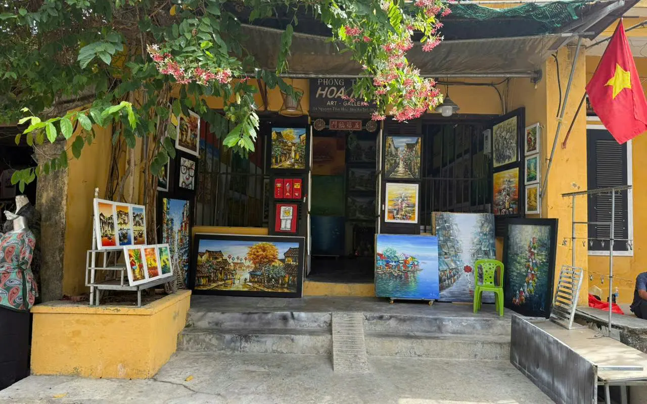 Art gallery entrance with colorful paintings displayed outside, surrounded by greenery, Vietnam flag visible.