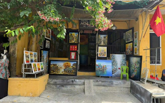 Art gallery entrance with colorful paintings displayed outside, surrounded by greenery, Vietnam flag visible.