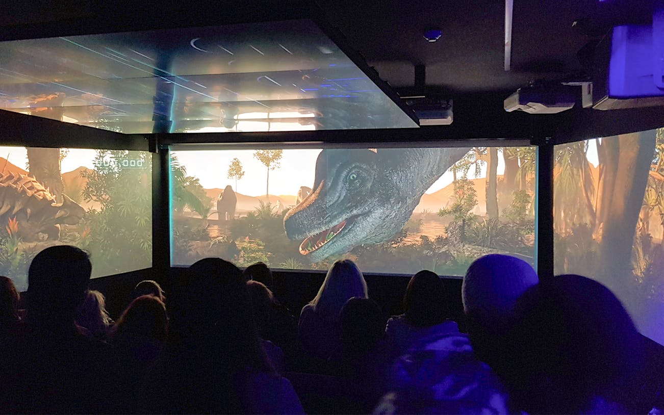 Audience watching an immersive underwater theatre experience with dinosaurs on screen.