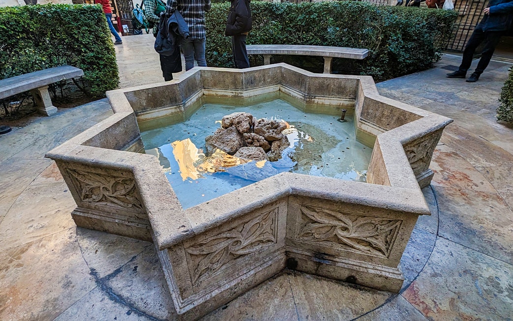 Stone fountain in the courtyard of San Nicolas Church, surrounded by benches and greenery.