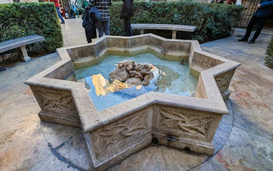 Stone fountain in the courtyard of San Nicolas Church, surrounded by benches and greenery.