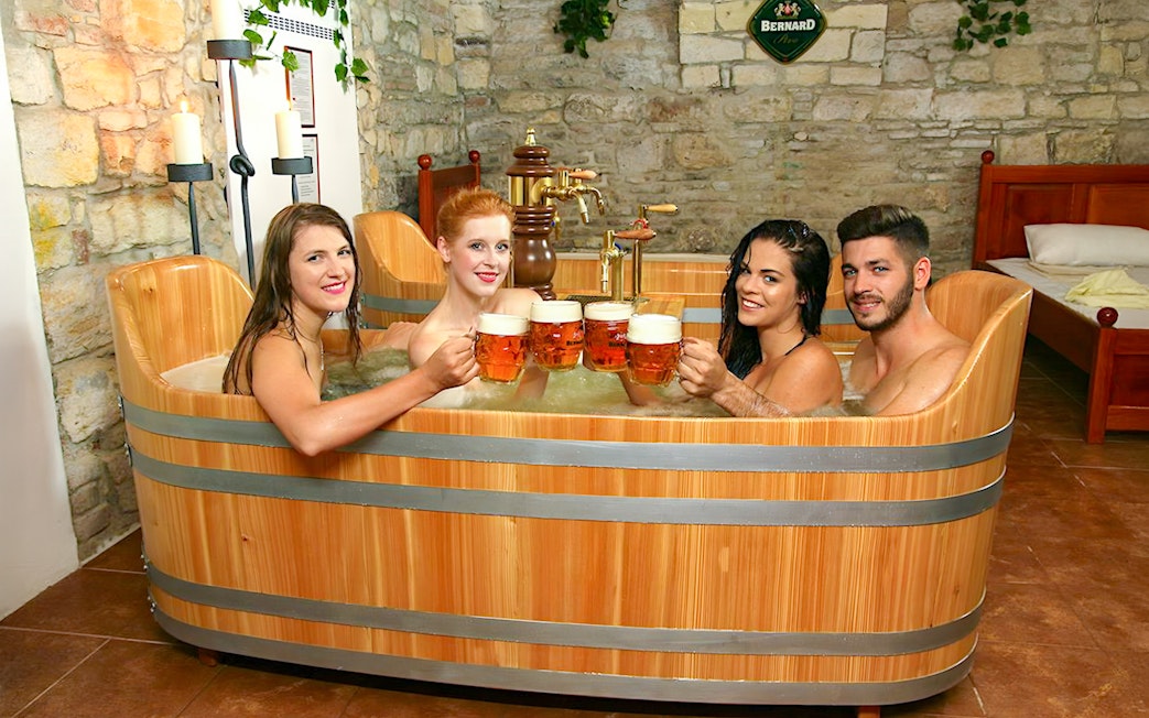 Guests enjoying a beer bath at Bernard Beer Spa, raising mugs in a wooden tub.
