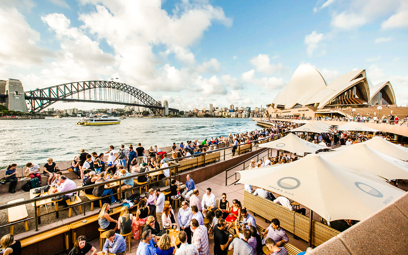 Crowd enjoying drinks at Opera Bar with Sydney Opera House and Harbour Bridge in view.