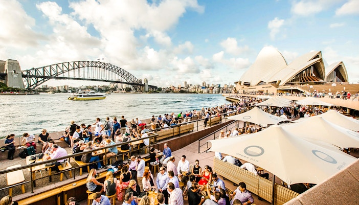 Crowd enjoying drinks at Opera Bar with Sydney Opera House and Harbour Bridge in view.