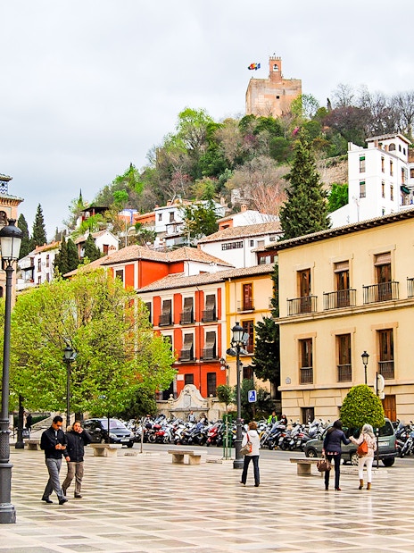 Plaza in Albaicin with view of Sacromonte, Granada, featuring historic buildings and people walking.
