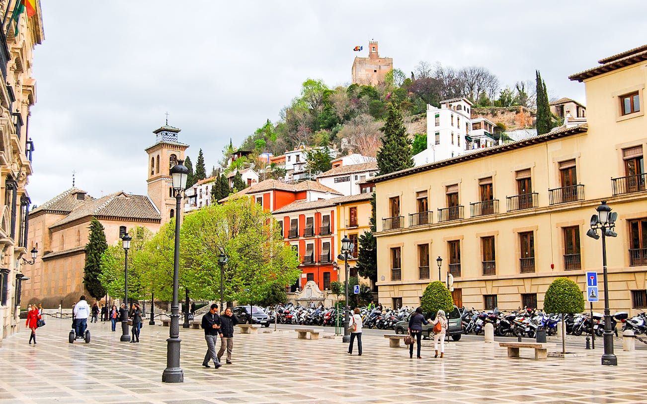 Plaza in Albaicin with view of Sacromonte, Granada, featuring historic buildings and people walking.