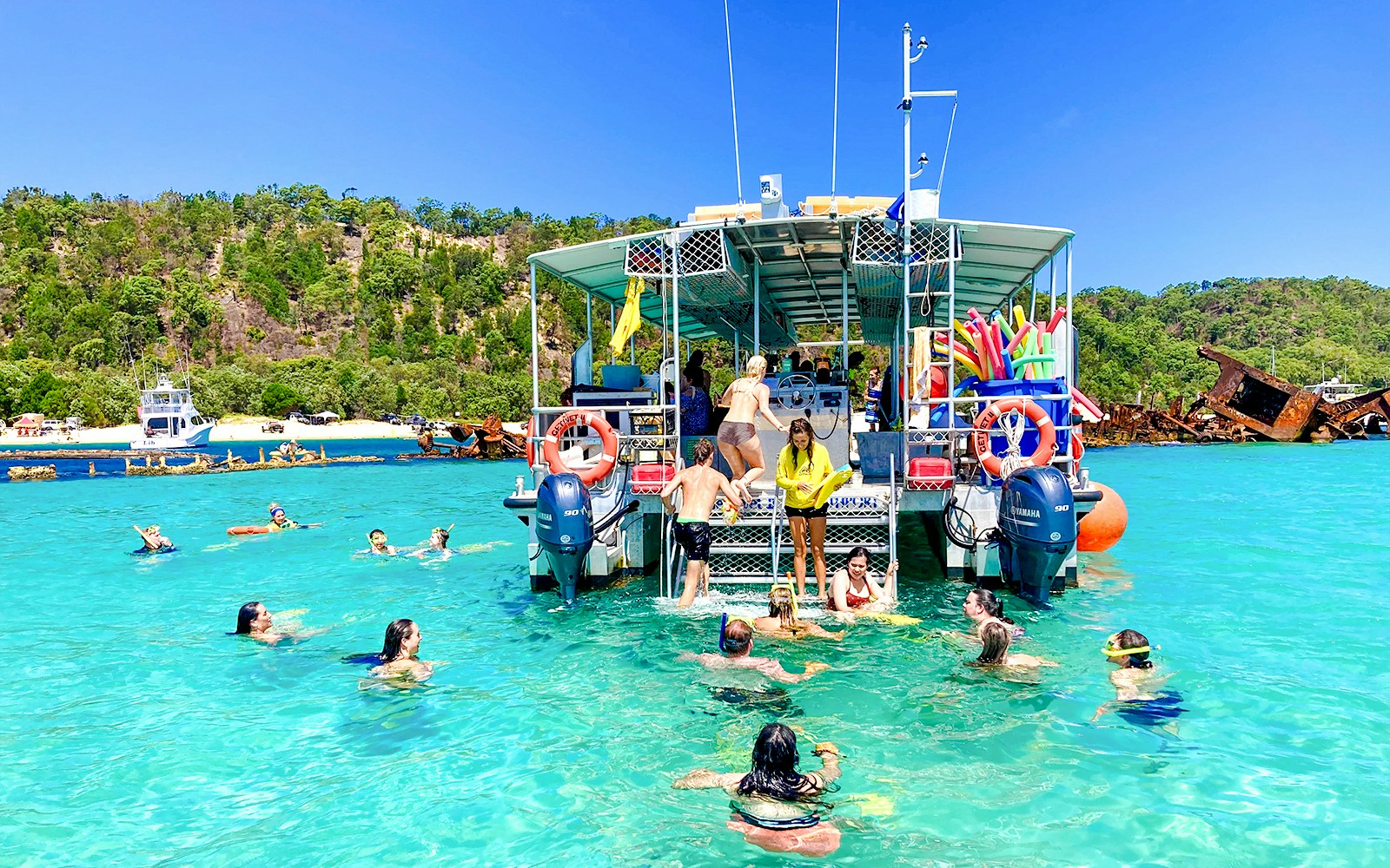 People swimming and snorkeling near a boat in Moreton Bay, with shipwrecks and lush greenery in the background.