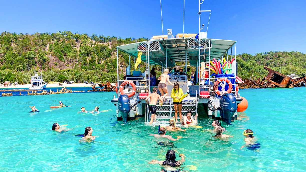 People swimming and snorkeling near a boat in Moreton Bay, with shipwrecks and lush greenery in the background.