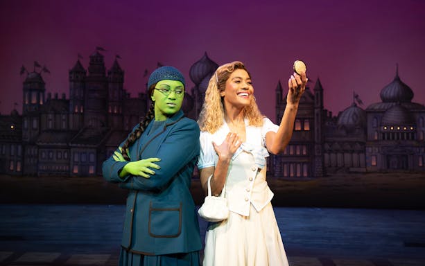 Two performers on stage in a scene from the musical Wicked, with a castle backdrop.
