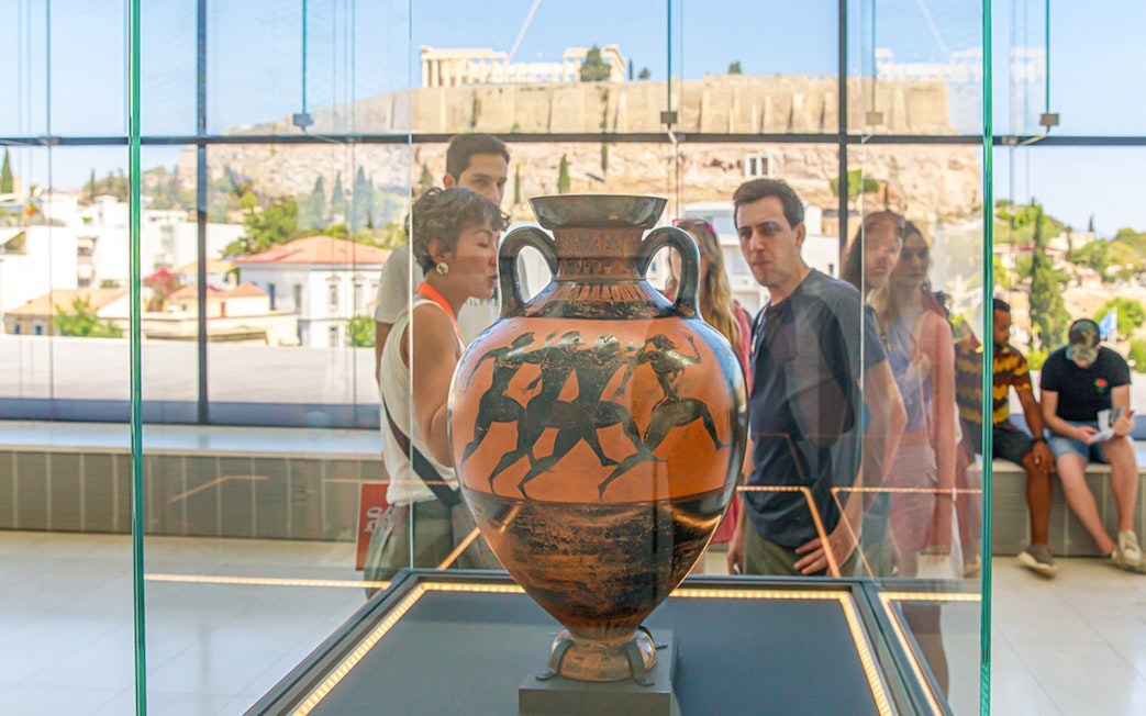 Visitors viewing ancient Greek vase at Athens Acropolis Museum with Parthenon in background.