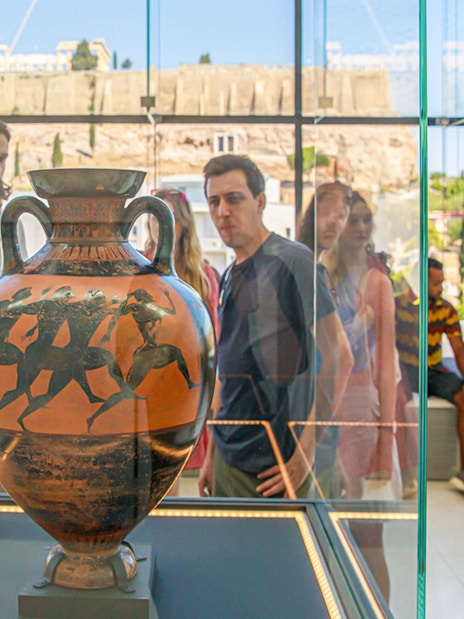 Visitors viewing ancient Greek vase at Athens Acropolis Museum with Parthenon in background.