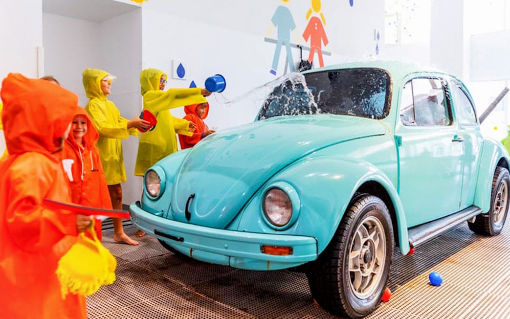 Children in raincoats playing with water near a vintage car at Water Gallery, Olioli Museum Dubai.
