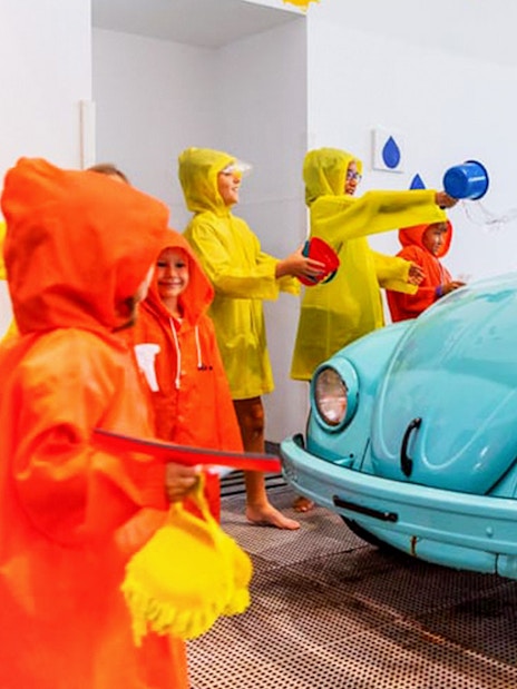 Children in raincoats playing with water near a vintage car at Water Gallery, Olioli Museum Dubai.