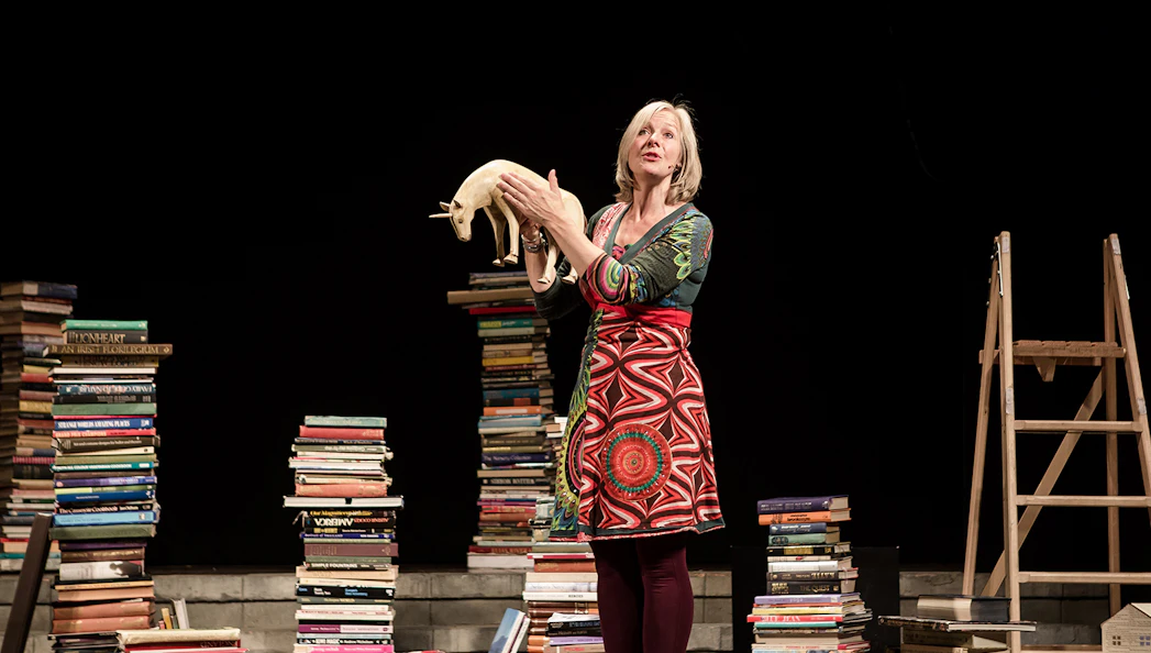 Performer holding a unicorn model surrounded by stacks of books on stage.