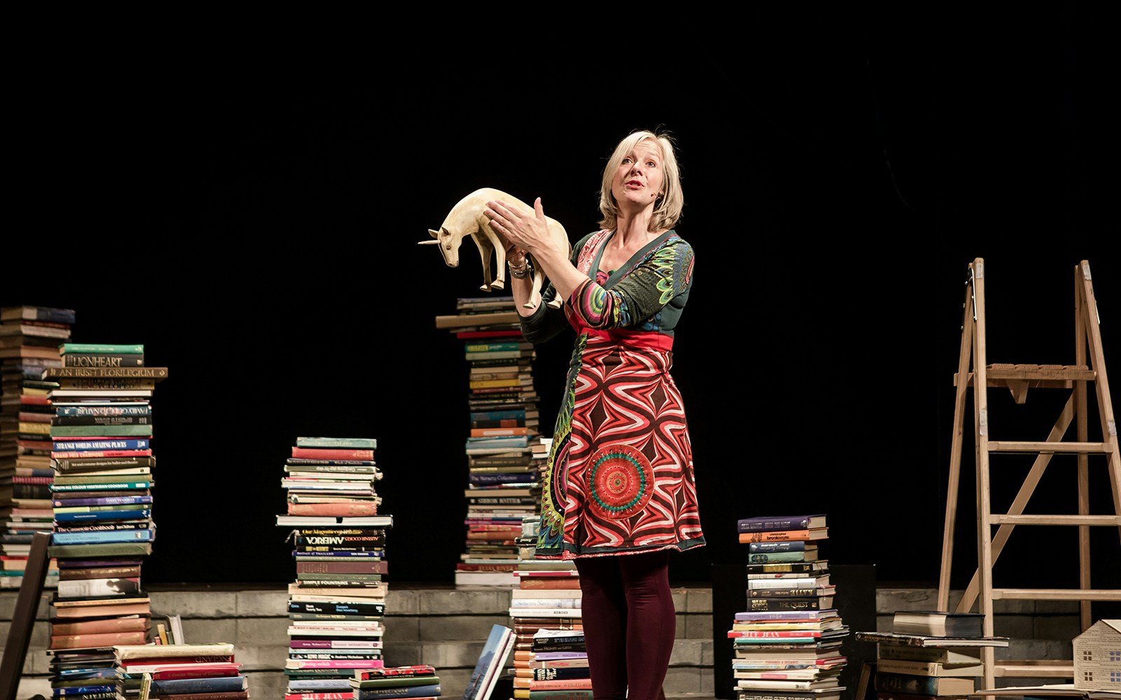 Performer holding a unicorn model surrounded by stacks of books on stage.