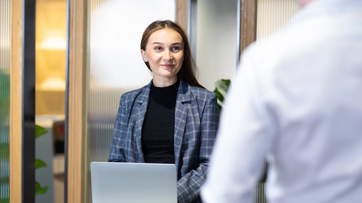 Woman welcoming visitor at a modern reception desk with a laptop.