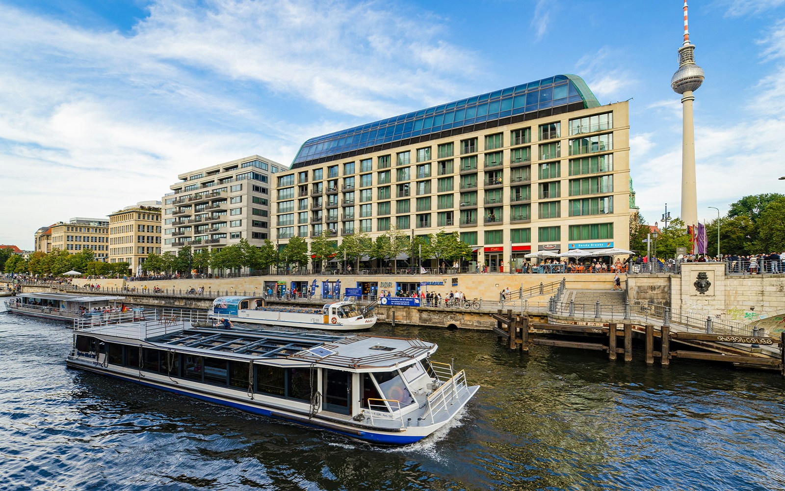 Die Spree in Berlin mit Ausflugsbooten, modernen Gebäuden und dem Fernsehturm im Hintergrund.