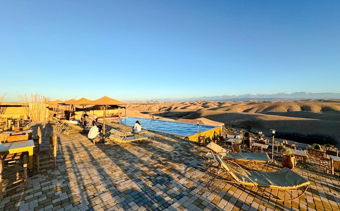 Desert camp with pool and seating area in Agafay, Marrakesh, overlooking dunes.