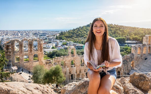 Woman sitting with a guidebook at the Acropolis, Athens, with ancient ruins in the background.