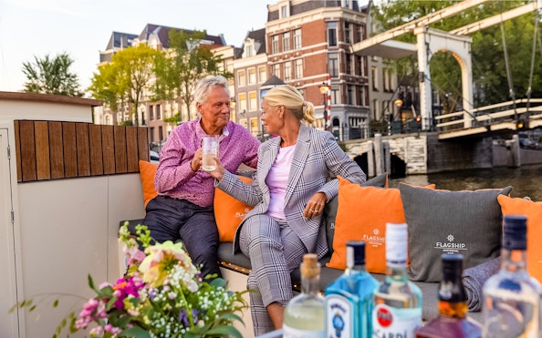 Couple enjoying drinks on a luxury boat in Amsterdam's canals with historic buildings in view.