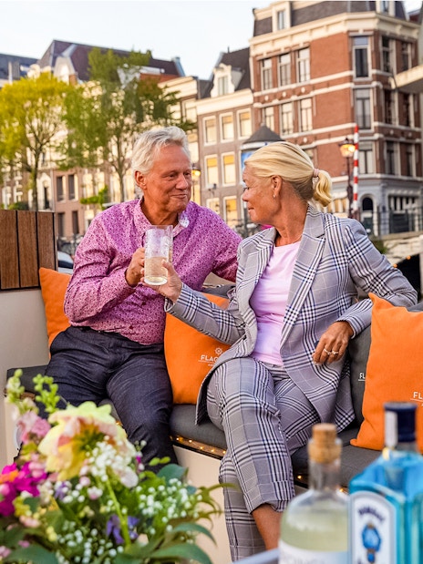 Couple enjoying drinks on a luxury boat in Amsterdam's canals with historic buildings in view.