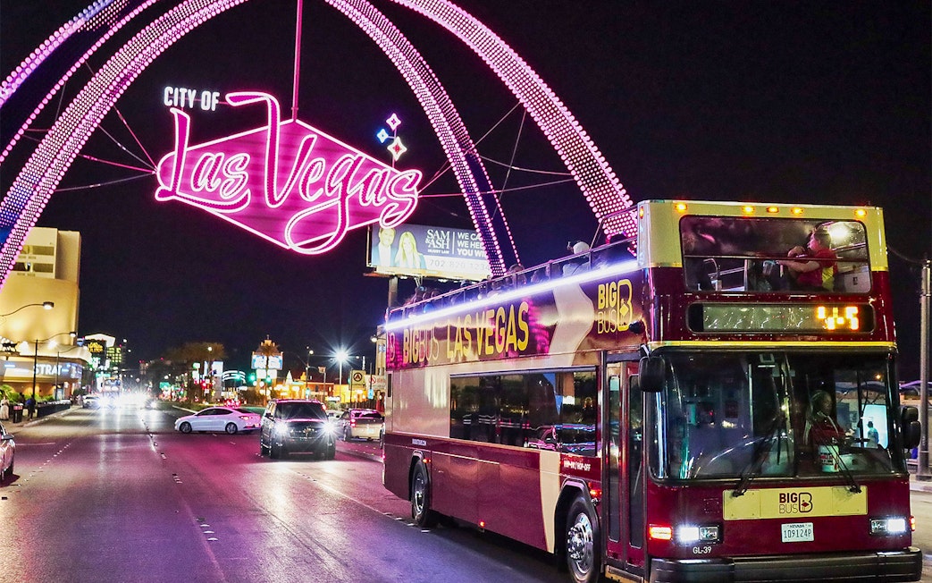 Big Bus tour in Las Vegas at night under neon city sign.