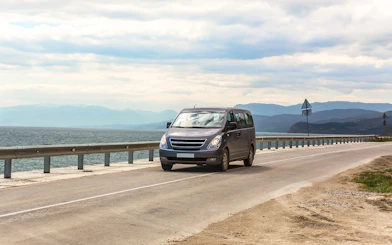 Minivan driving on coastal highway with sea and mountains in the background.