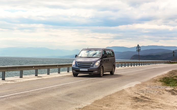 Minivan driving on coastal highway with sea and mountains in the background.
