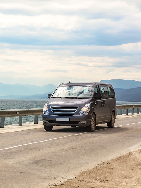 Minivan driving on coastal highway with sea and mountains in the background.
