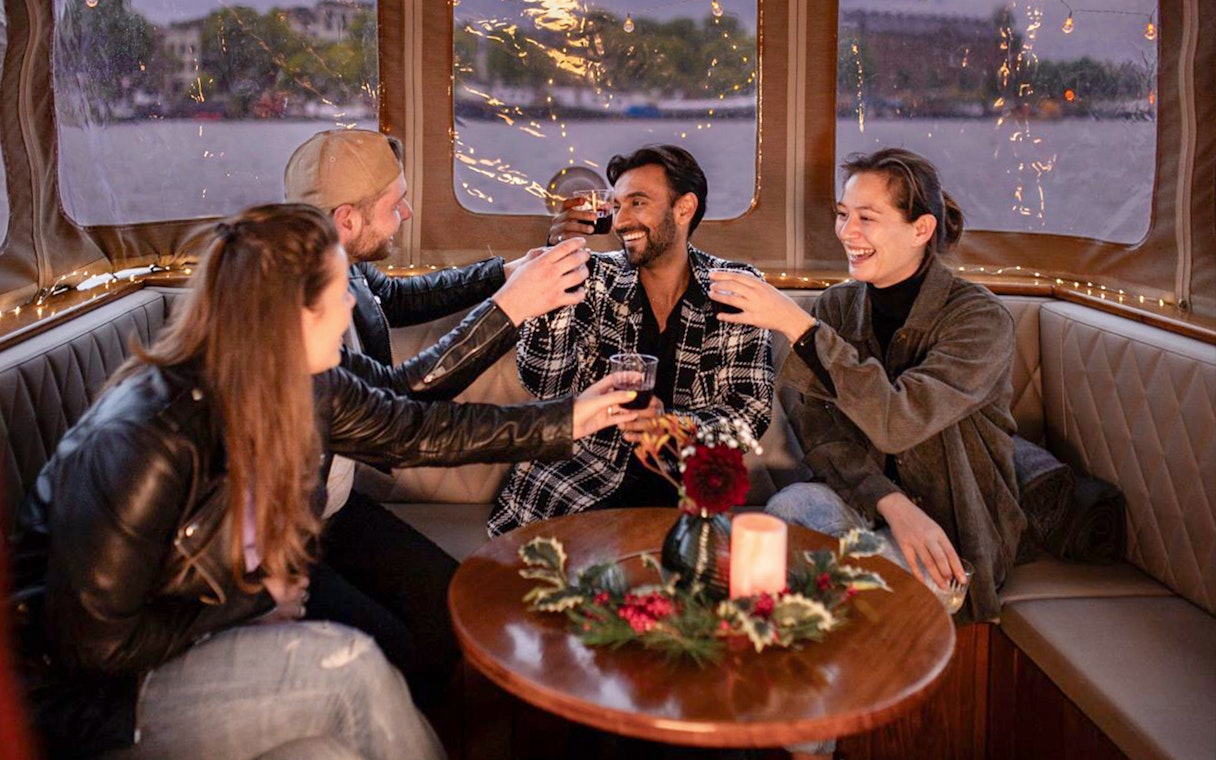 Friends toasting on a cozy Amsterdam Light Festival cruise with festive decor.