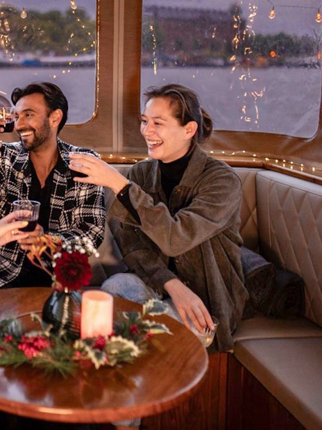 Friends toasting on a cozy Amsterdam Light Festival cruise with festive decor.