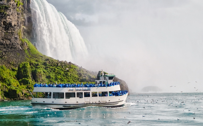 Tourists on a boat near the base of Niagara Falls, surrounded by mist and birds.
