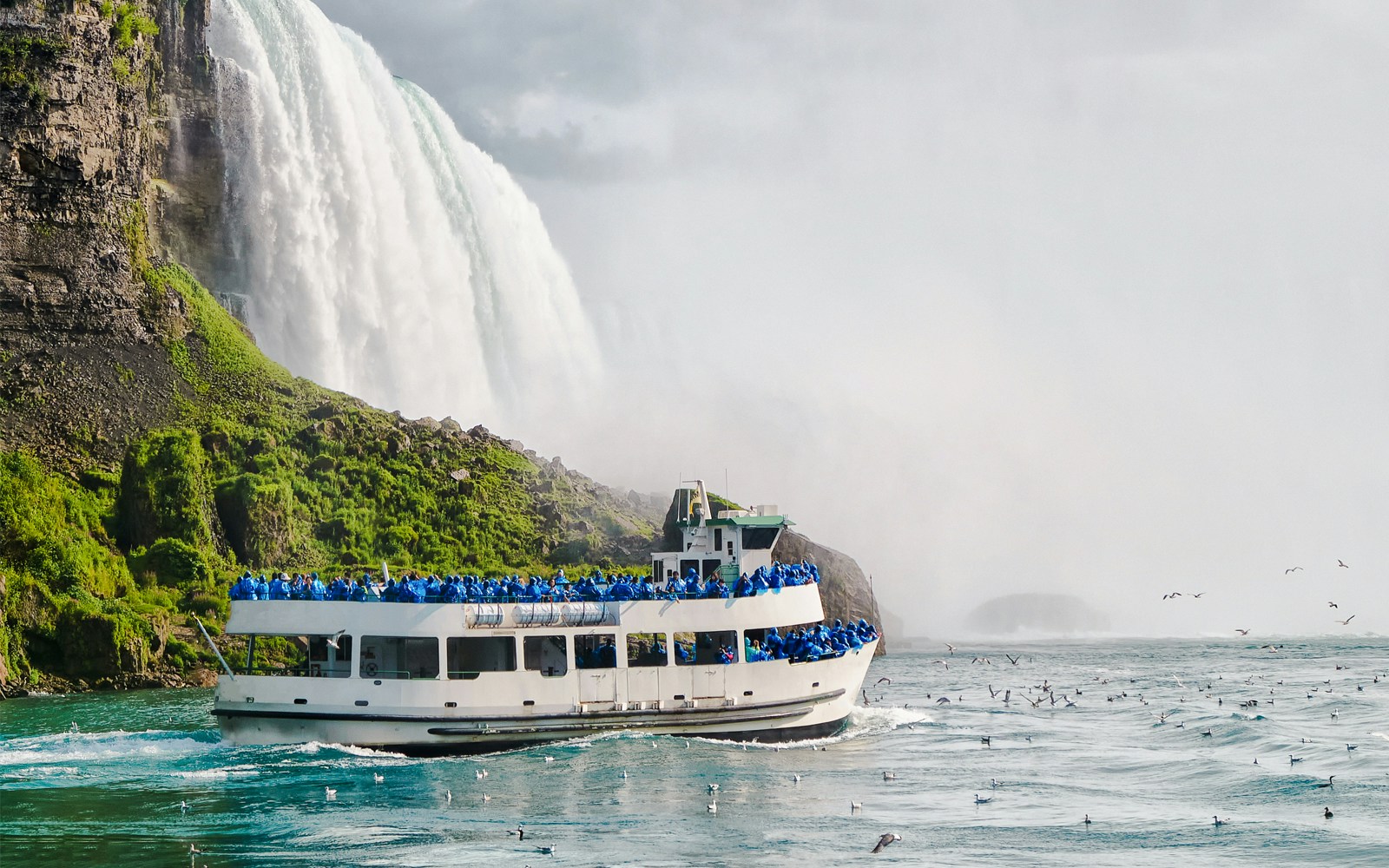 Tourists on a boat near the base of Niagara Falls, surrounded by mist and birds.