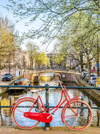 Red bicycle on a bridge over an Amsterdam canal with trees and buildings in the background.