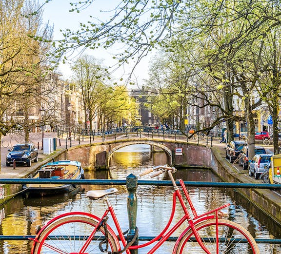 Red bicycle on a bridge over an Amsterdam canal with trees and buildings in the background.