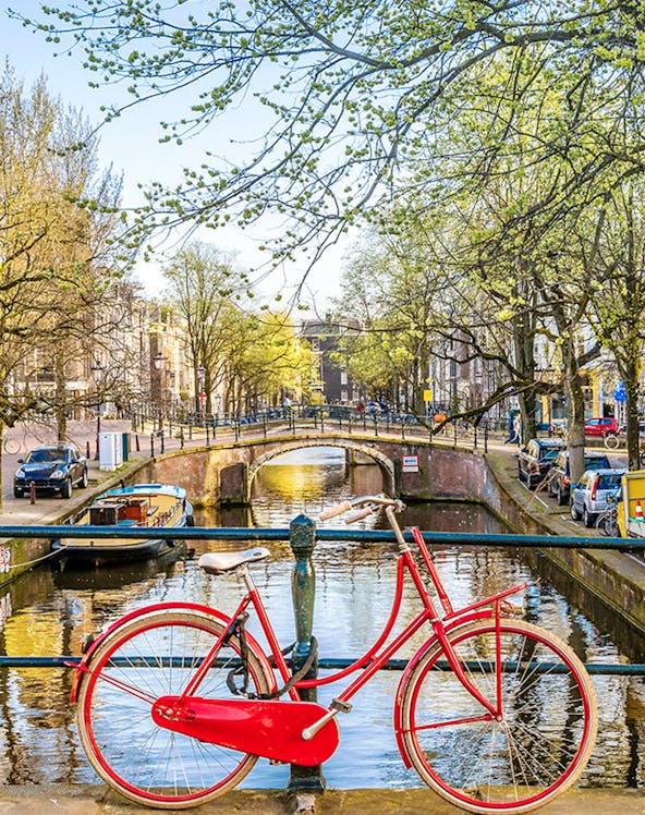 Red bicycle on a bridge over an Amsterdam canal with trees and buildings in the background.