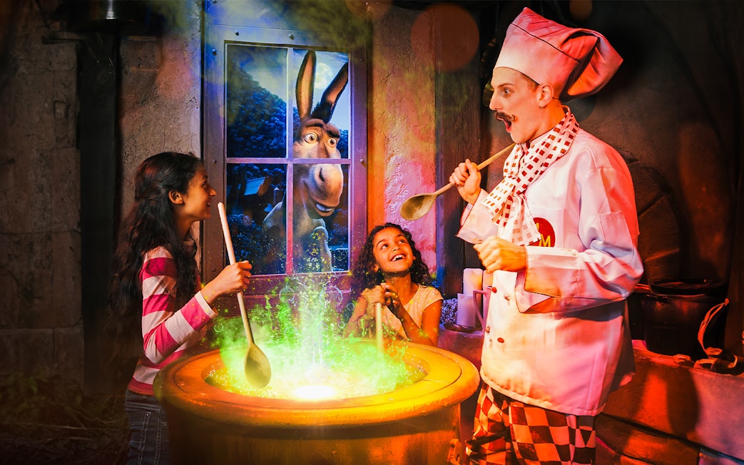 Children stirring a cauldron with a chef at Shrek’s Adventure! London, Donkey visible through window.