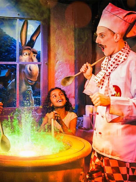 Children stirring a cauldron with a chef at Shrek’s Adventure! London, Donkey visible through window.