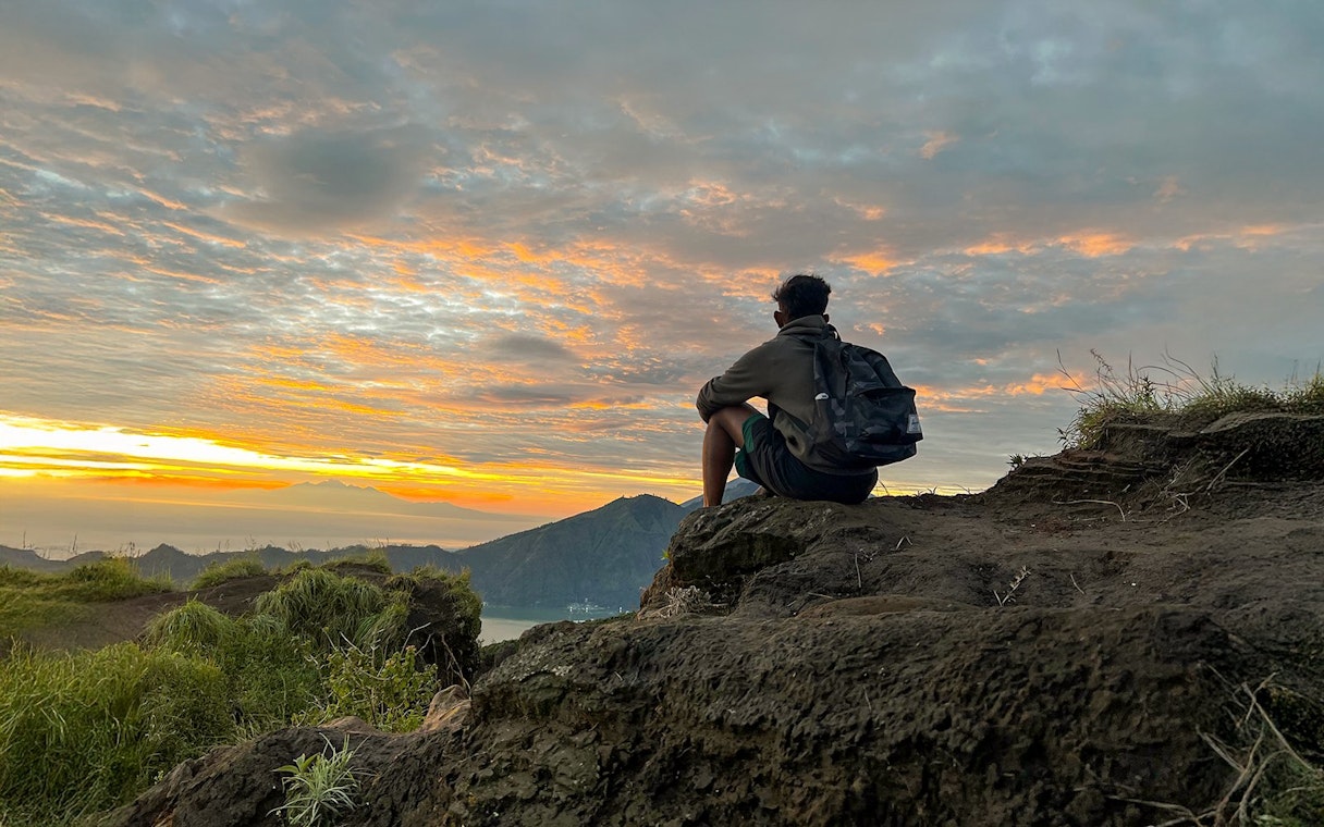 Person sitting on rocky ledge watching sunrise at Mount Batur, Bali.
