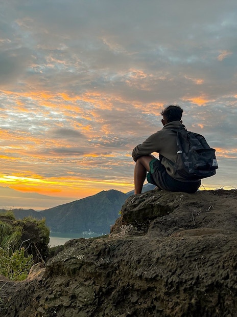 Person sitting on rocky ledge watching sunrise at Mount Batur, Bali.