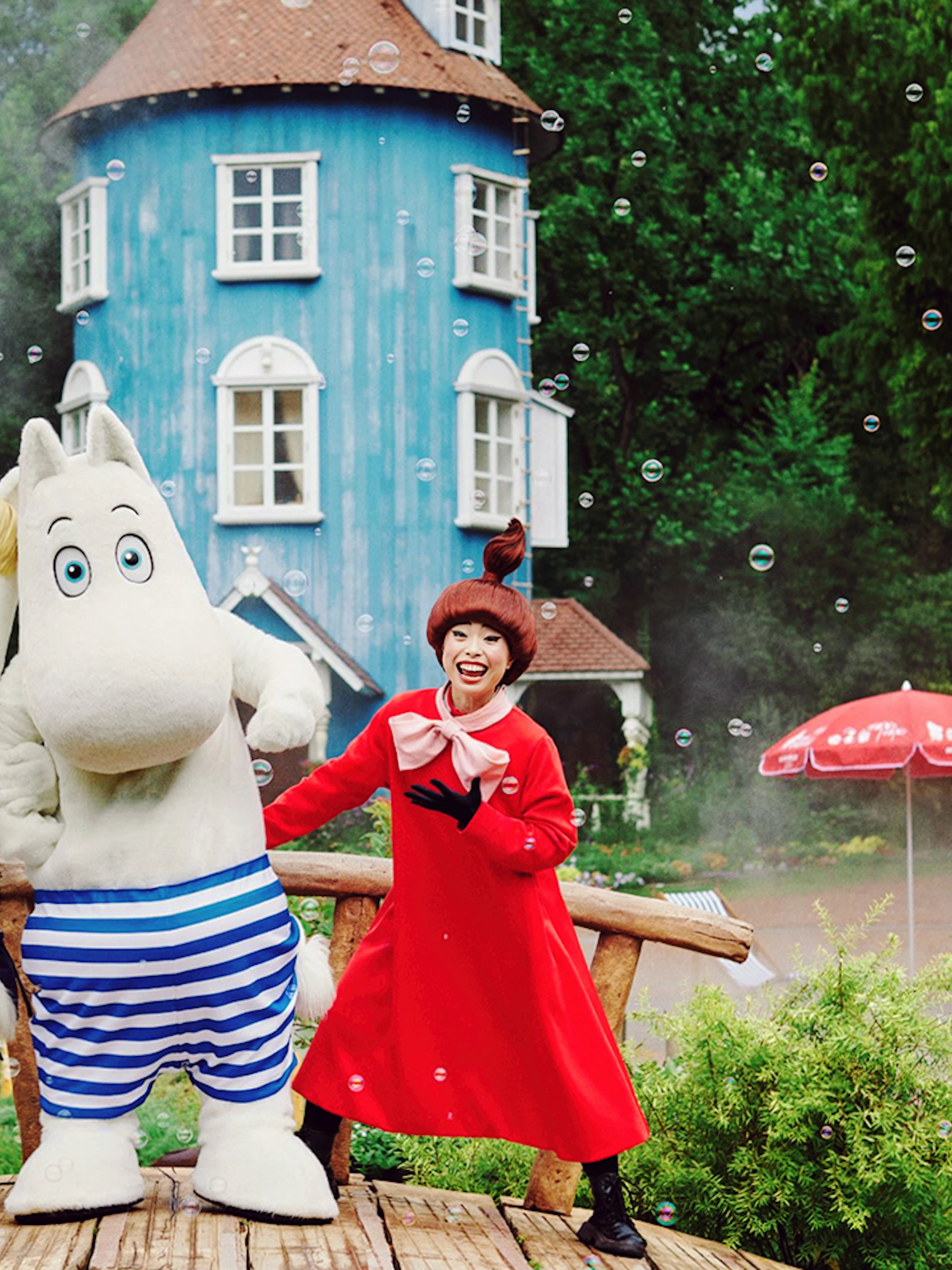 Moomin characters and a performer in front of a blue house at Moominvalley Park.