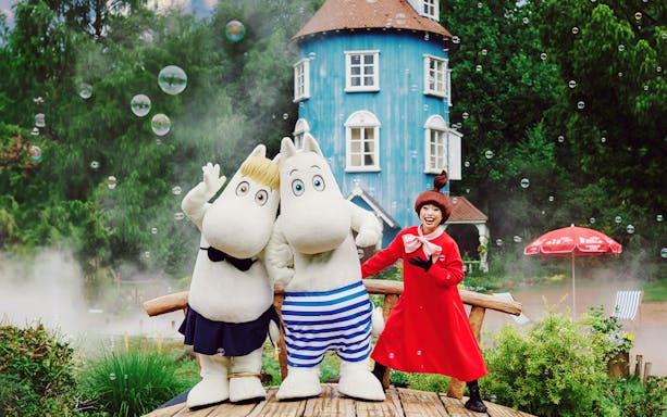 Moomin characters and a performer in front of a blue house at Moominvalley Park.