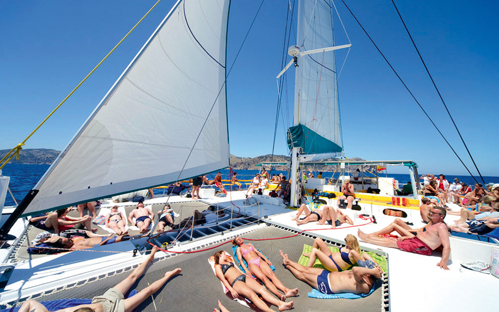 People relaxing on a catamaran deck during a trip in Alcudia, with sails and sea in the background.