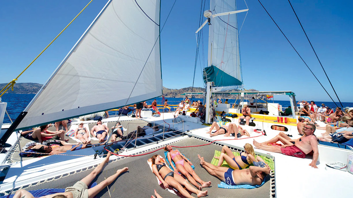 People relaxing on a catamaran deck during a trip in Alcudia, with sails and sea in the background.