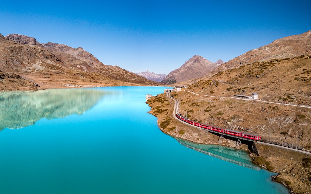 Bernina Express train crossing a bridge over a turquoise lake in the Swiss Alps.