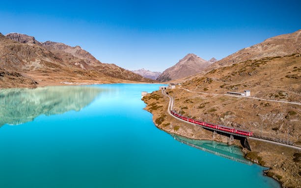 Bernina Express train crossing a bridge over a turquoise lake in the Swiss Alps.