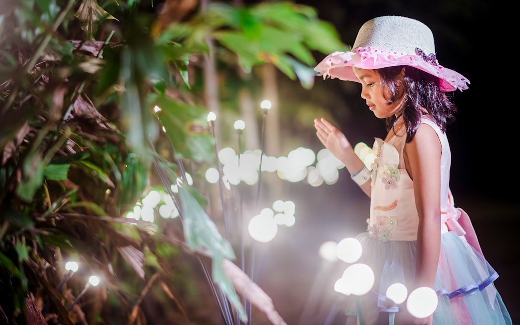 Little girl in a hat admiring glowing lights in a garden setting.