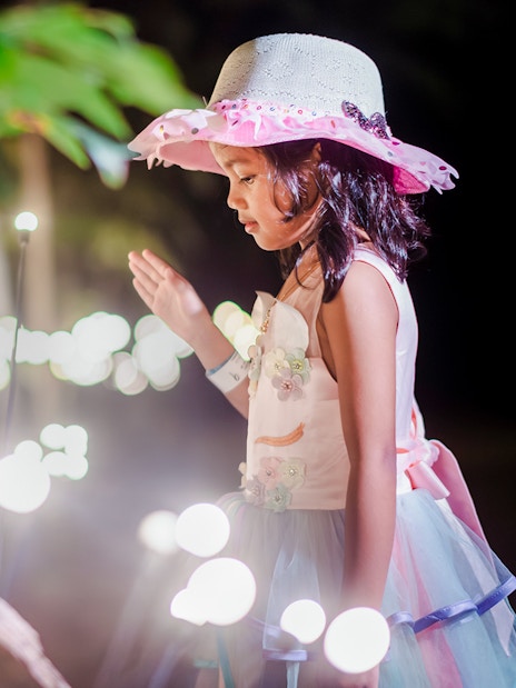 Little girl in a hat admiring glowing lights in a garden setting.