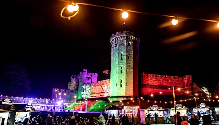 Warwick Castle illuminated at night during the Light Trail event, with visitors exploring festive stalls.